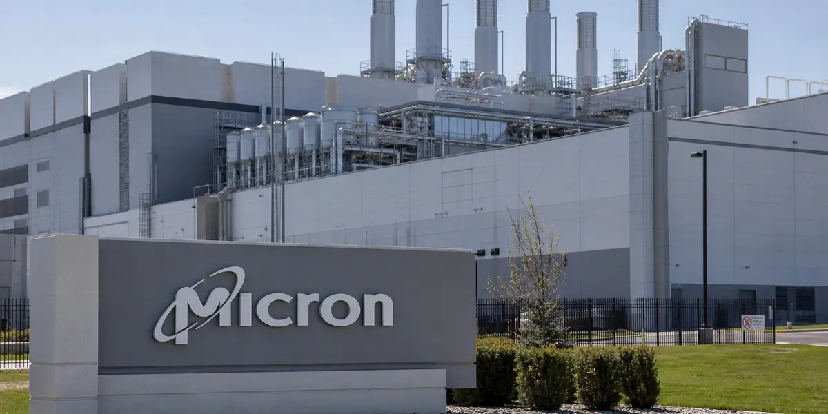 대형 반도체 생산공장 외관을 담은 사진으로, 클린룸 공조 설비가 얹힌 흰색 타워 구조물과 회사 로고가 새겨진 표지판이 전면에 크게 보이고, 뒤편으로 넓은 부지와 맑은 하늘이 펼쳐진 산업단지 전경의 모습